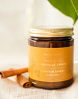 Gros plan sur bougie Pumpkin Spice dans bocal en verre ambré avec couvercle doré, bâtons de cannelle et tissu en lin naturel
