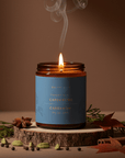 Gros plan sur bougie Cardamome en verre ambré avec flamme dorée et étiquette botanique sur tranche de bois rustique