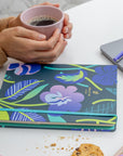 Hands holding a pale pink coffee cup beside vibrant floral-patterned undated planners on a clean white table