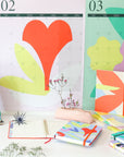 Agenda non daté Bouquet posé sur un bureau blanc entouré de calendriers muraux colorés et de feuillages verts dans un espace lumineux