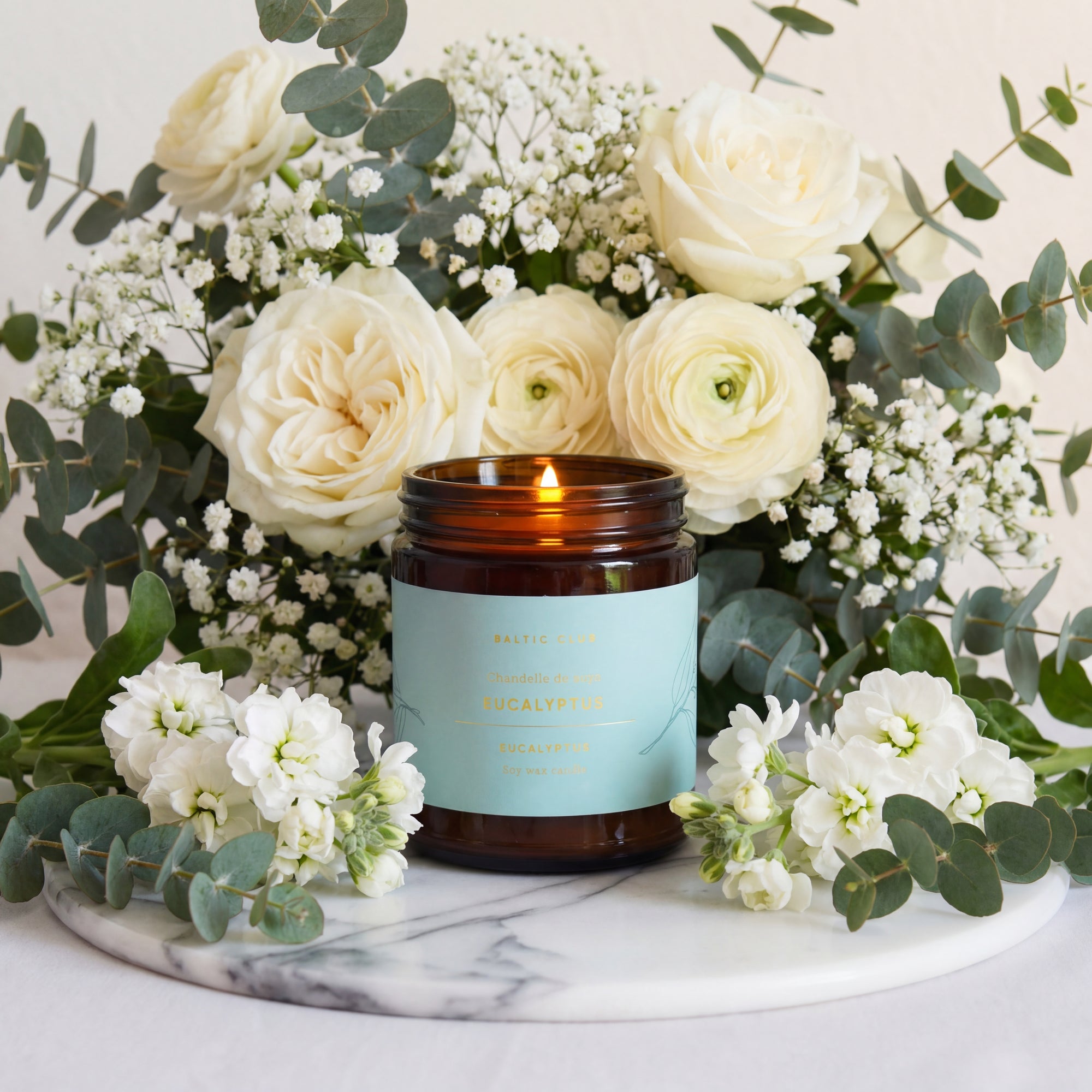 Bougie en verre ambré allumée parfum eucalyptus, entourée de fleurs blanches et verdure sur plateau marbré, ambiance sereine.