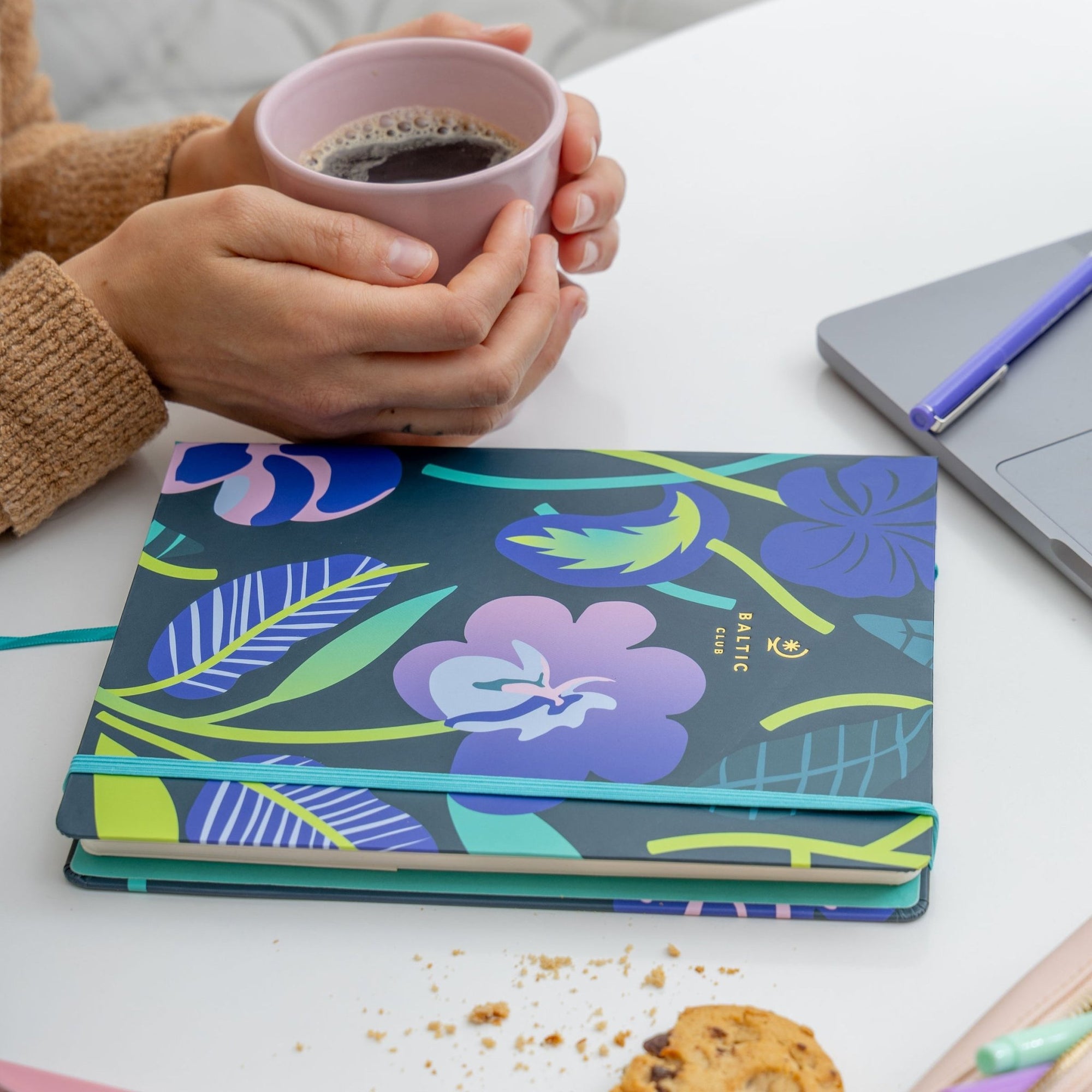 Hands holding a pale pink coffee cup beside vibrant floral-patterned undated planners on a clean white table