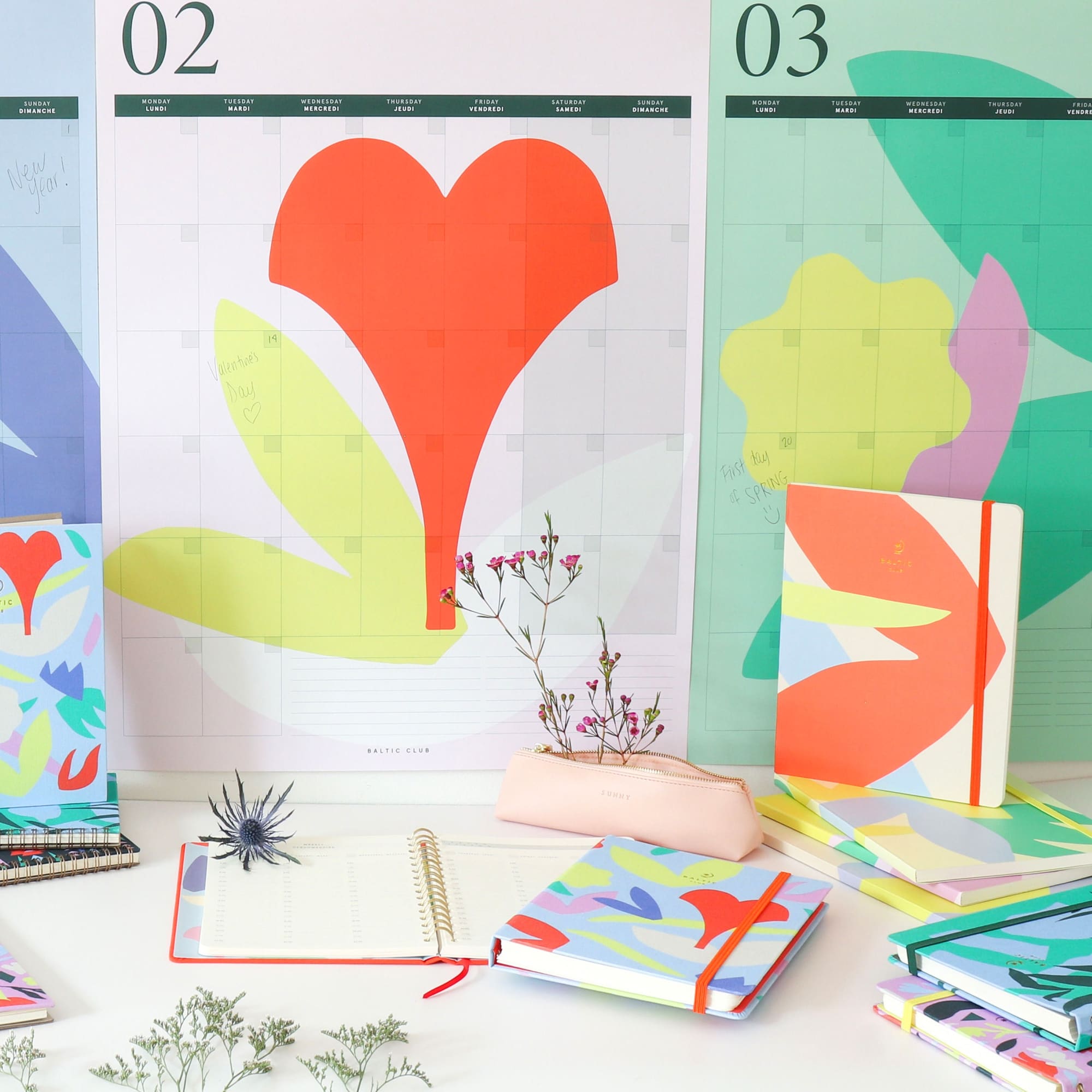 Agenda non daté Bouquet posé sur un bureau blanc entouré de calendriers muraux colorés et de feuillages verts dans un espace lumineux