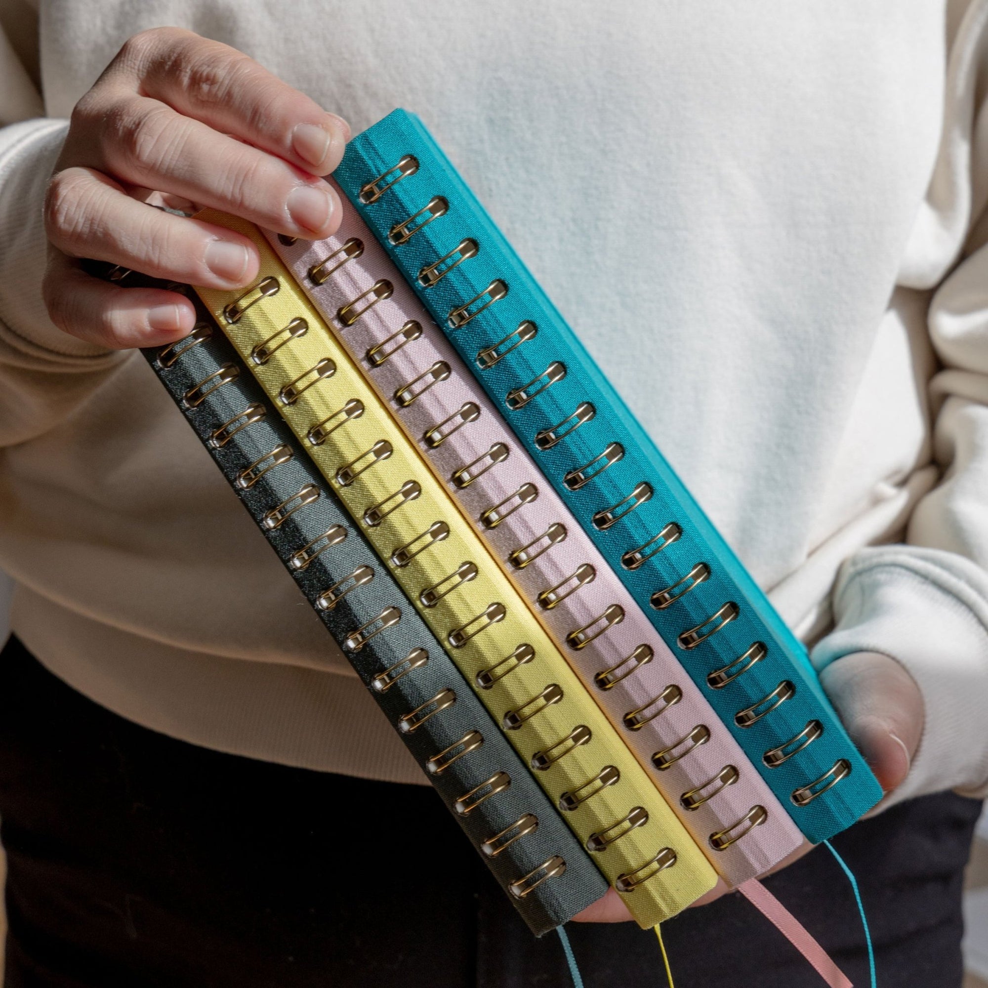 Mains tenant quatre carnets spirales aux couleurs pastel et reliures dorées sur fond flou doux