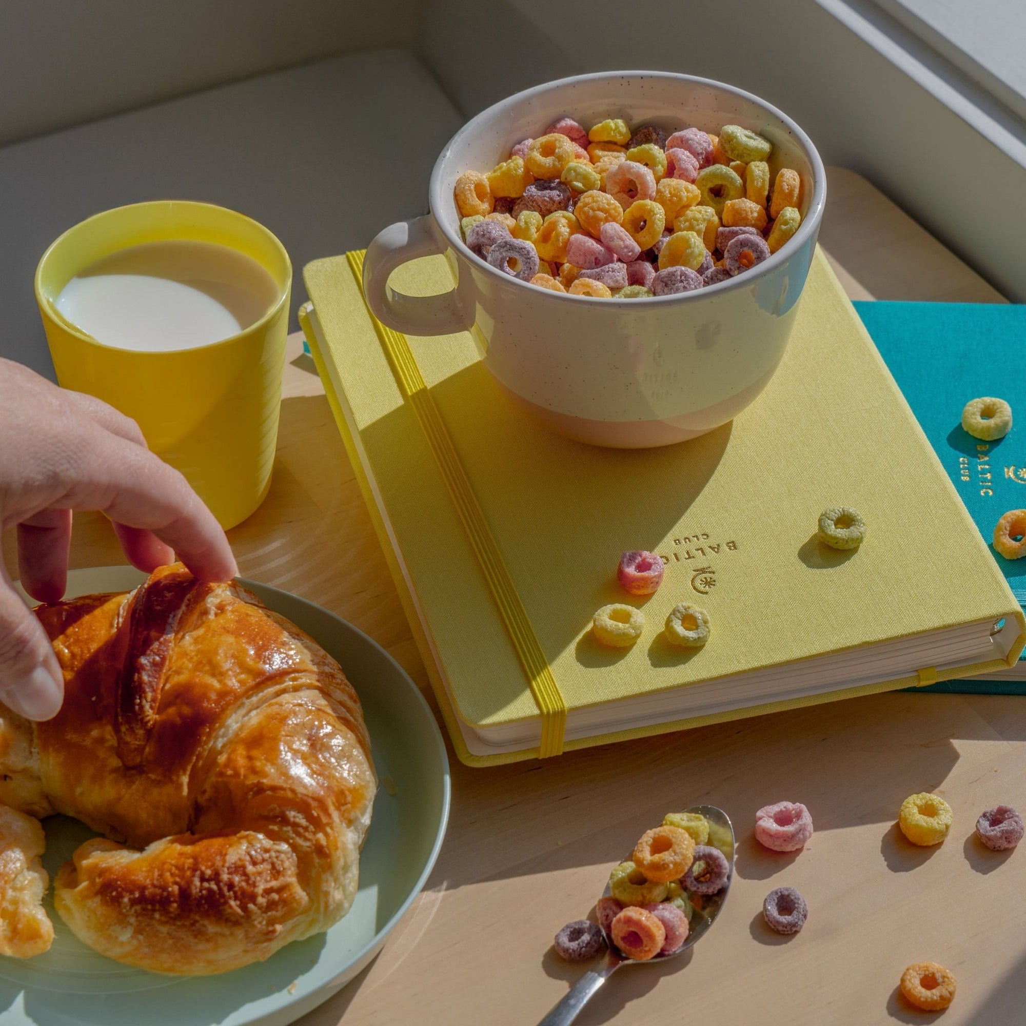 Scène de petit-déjeuner lumineuse avec bol de céréales colorées, croissant sur assiette bleu pâle et carnet jaune sur table en bois