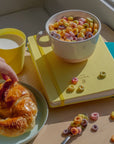 Scène de petit-déjeuner lumineuse avec bol de céréales colorées, croissant sur assiette bleu pâle et carnet jaune sur table en bois