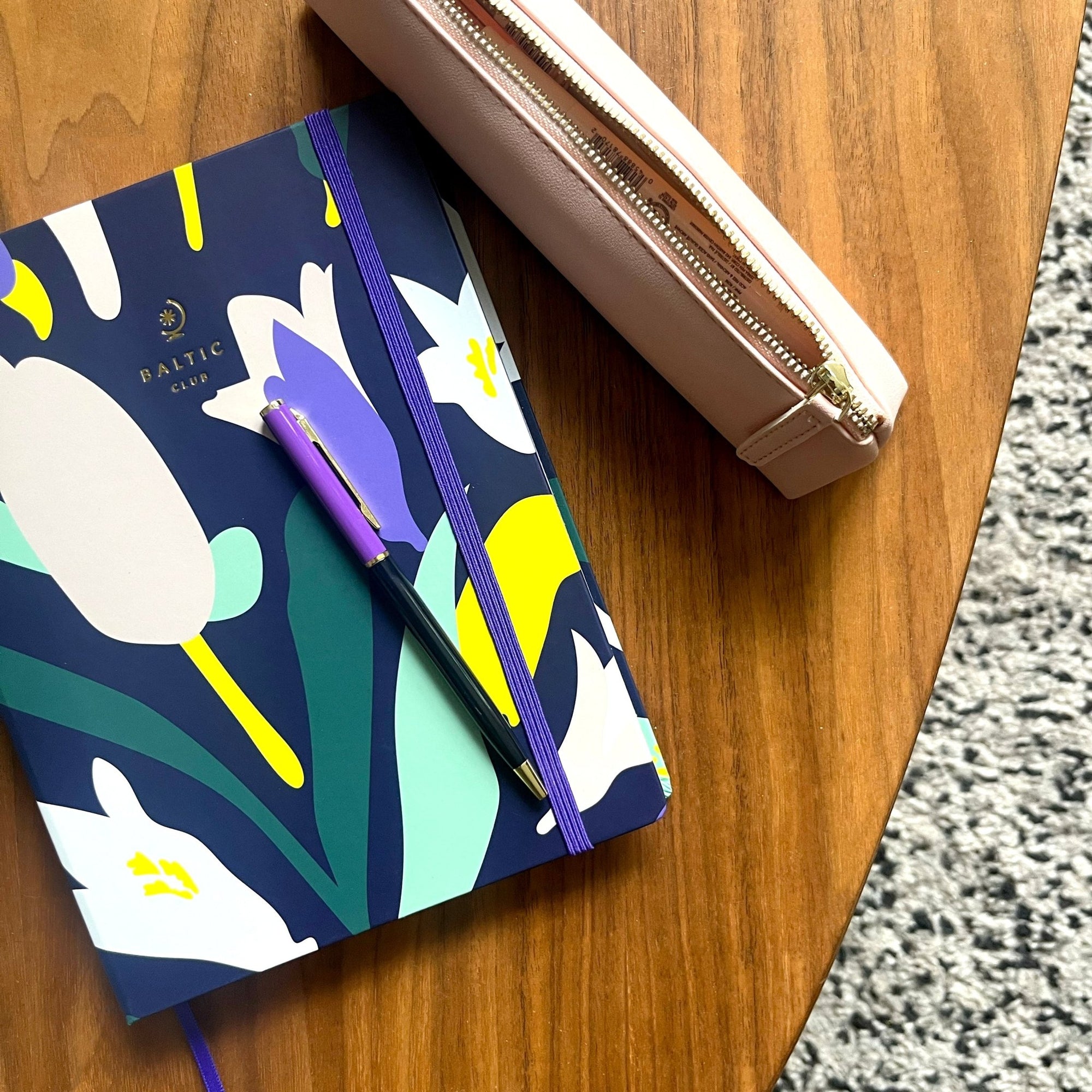 Vue en plongée oblique du carnet à dessins Hypnose bleu marine mat, motifs floraux vifs, logo doré, élastique violet et stylo assorti