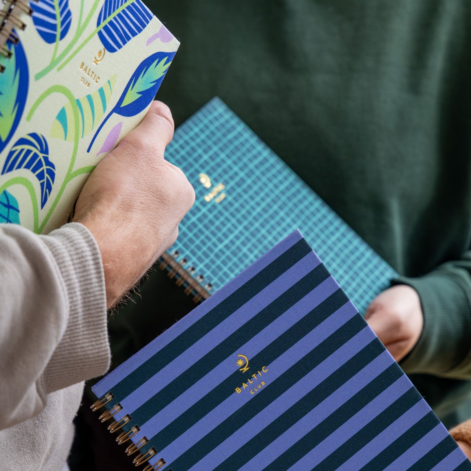 Trois carnets à spirale Baltic Club aux couvertures florales et géométriques tenus par des mains sous une lumière naturelle douce