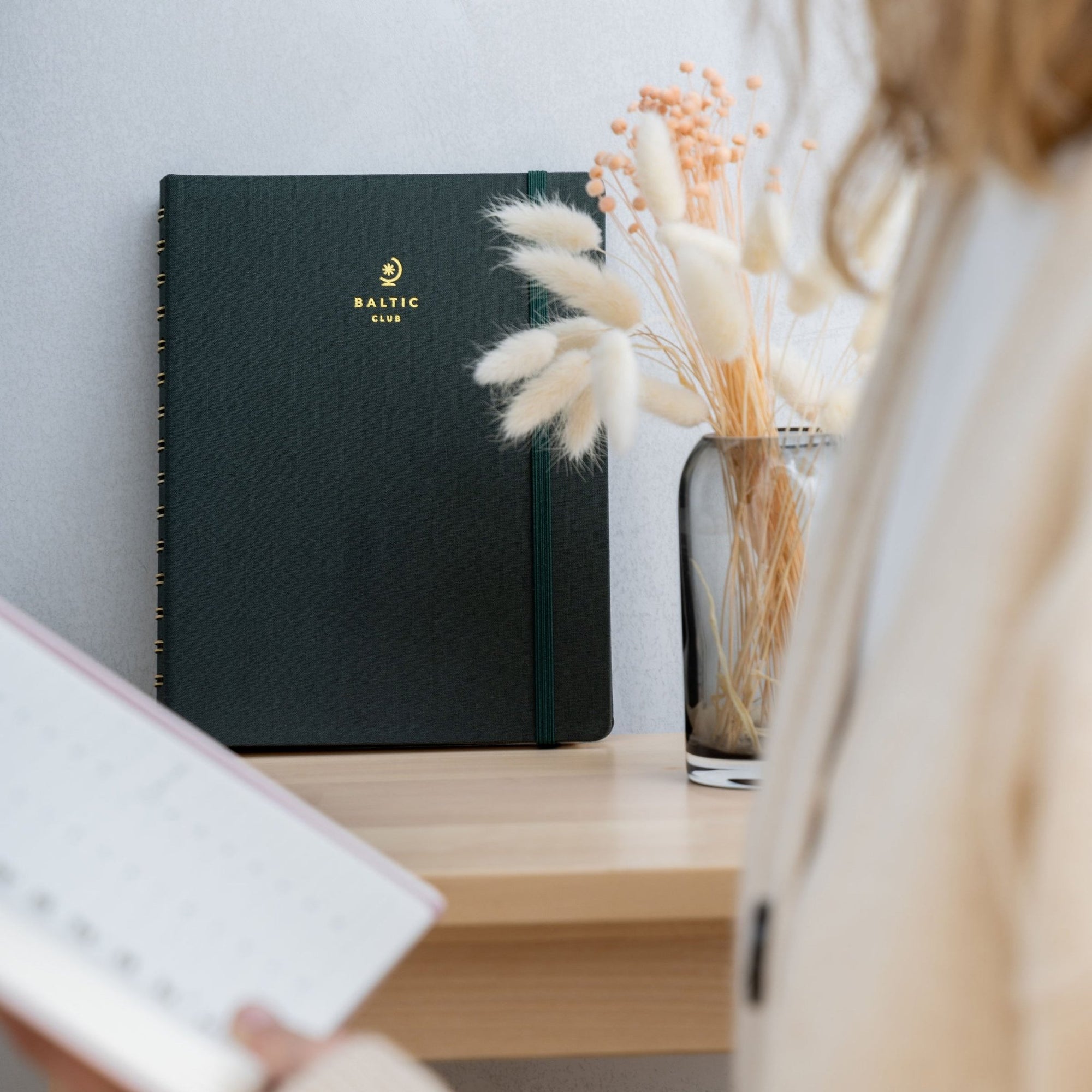Agenda vert foncé élégant Baltic Club avec logo doré sur table en bois, fleurs séchées et lumière naturelle douce