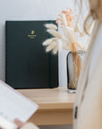 Agenda vert foncé élégant Baltic Club avec logo doré sur table en bois, fleurs séchées et lumière naturelle douce