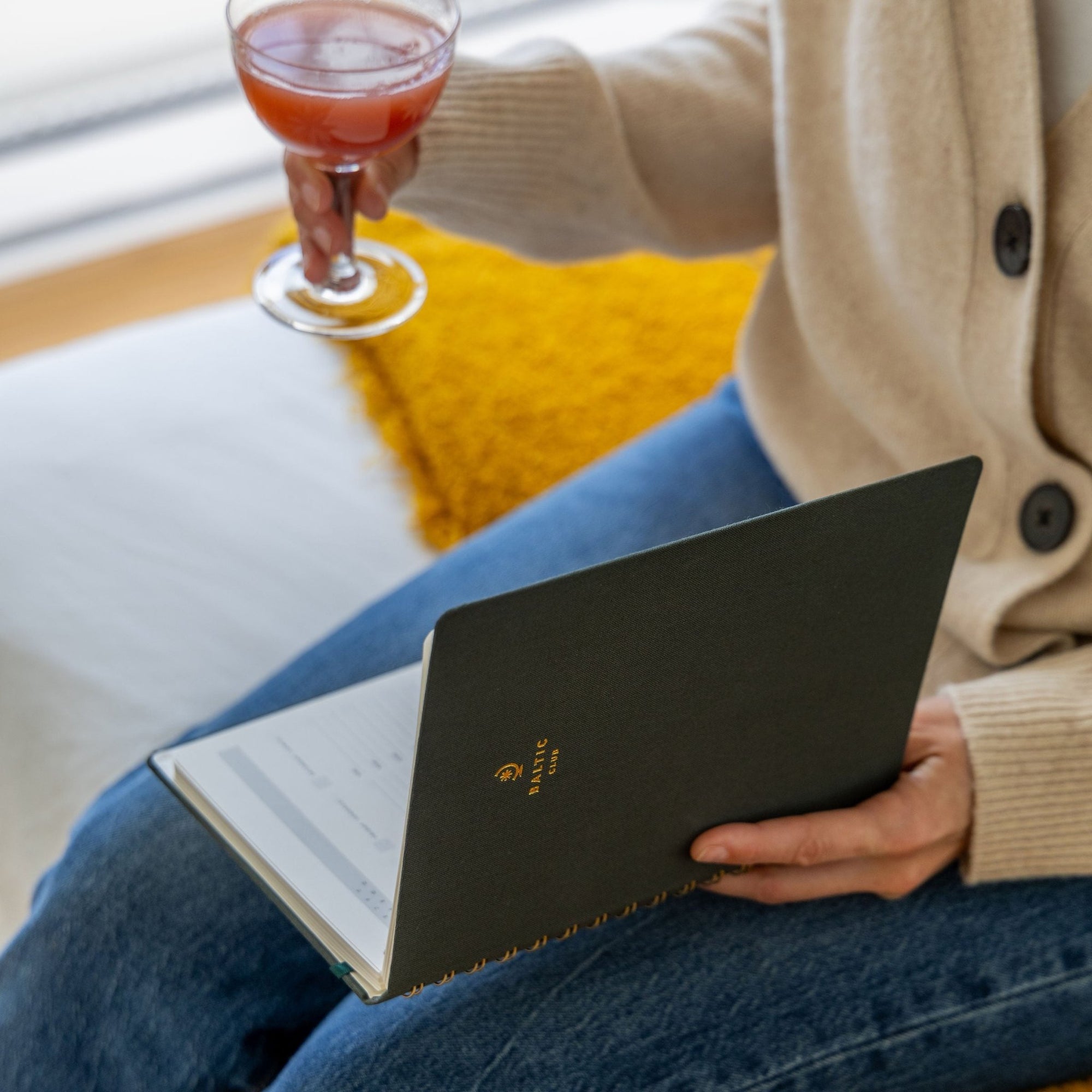 Scène chaleureuse avec un carnet spirale vert foncé Baltic Club et un cocktail rose, textures douces et lumière tamisée