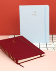 The Blue Ash Cloth-covered Undated Planner on a table next to a burgundy-covered one
