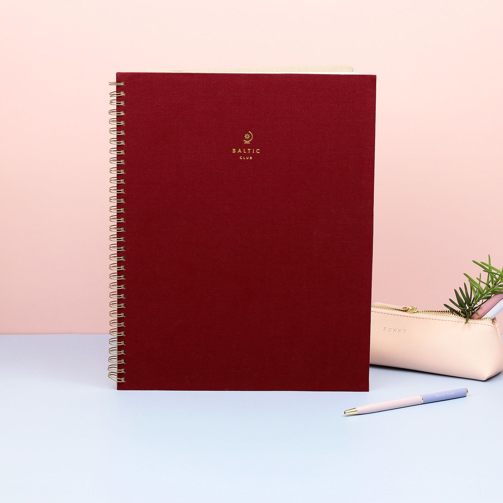 The large burgundy cloth-covered spiral notebook by the Baltic Club standing in front of pink decor